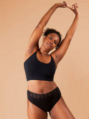A Black woman standing and looking directly at the camera with a slight smile, raising her hands in the air as if stretching. She is wearing black period pants with floral lace trim and a cropped bra.